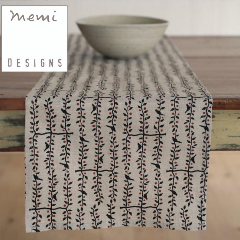 May include: A beige table runner with a repeating pattern of black birds and red berries on vines. The text "memi DESIGNS" is in a white box in the upper left corner. A light-colored bowl sits on the table runner.