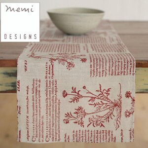May include: A beige table runner with a red botanical print and text. The runner features a repeating pattern of floral illustrations and script. The "memi DESIGNS" logo is in the upper left corner. A light-colored bowl sits on the runner.