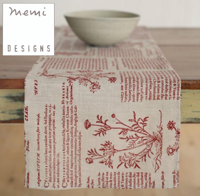 May include: A beige table runner with a red botanical print and text. The runner features a repeating pattern of floral illustrations and script. The "memi DESIGNS" logo is in the upper left corner. A light-colored bowl sits on the runner.