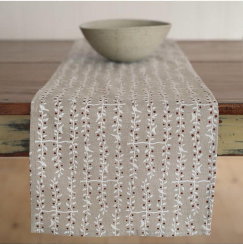 May include: A beige table runner with a repeating pattern of white branches and red berries. A light-colored bowl sits on the runner. The runner is made of a natural-looking fabric and is placed on a wooden table.