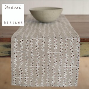 May include: Beige table runner with a white botanical pattern featuring small birds and berries. The runner is made of a natural-looking fabric and is topped with a light-colored bowl. The text "MEMI DESIGNS" is in the upper left corner.