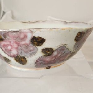 May include: A white ceramic bowl with a scalloped rim, adorned with pink and brown floral designs. The bowl has a rustic, handmade look, ideal for serving food or as a decorative item.