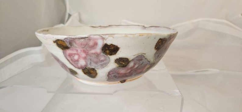 May include: A white ceramic bowl with a scalloped rim, adorned with pink and brown floral designs. The bowl has a rustic, handmade look, ideal for serving food or as a decorative item.