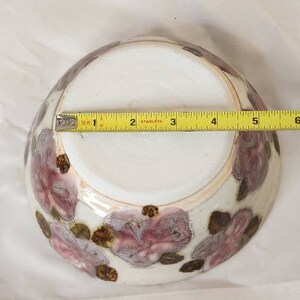 May include: A white ceramic bowl with a floral design in shades of purple and brown. The bowl is approximately 15 cm in diameter. The floral pattern is hand-painted, adding a unique touch to the piece.