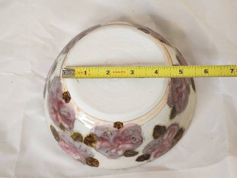 May include: A white ceramic bowl with a floral design in shades of purple and brown. The bowl is approximately 15 cm in diameter. The floral pattern is hand-painted, adding a unique touch to the piece.