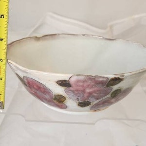 May include: A white ceramic bowl with a floral design. The bowl has a scalloped rim and is decorated with pink and brown flower motifs. The bowl is approximately 7.6 cm in height and 15.2 cm in diameter.