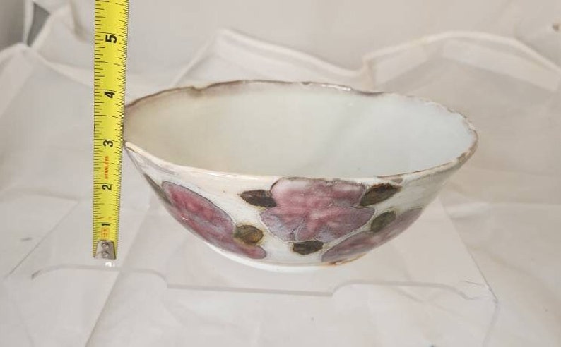 May include: A white ceramic bowl with a floral design. The bowl has a scalloped rim and is decorated with pink and brown flower motifs. The bowl is approximately 7.6 cm in height and 15.2 cm in diameter.