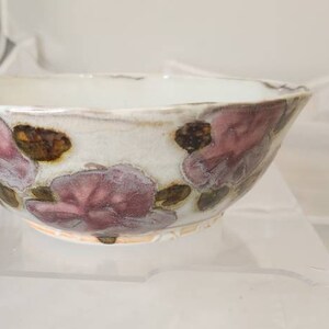 May include: A ceramic bowl with a white interior and exterior, decorated with pink and brown floral patterns. The bowl has a slightly uneven rim, giving it a rustic, handmade look. It is likely used for serving food.