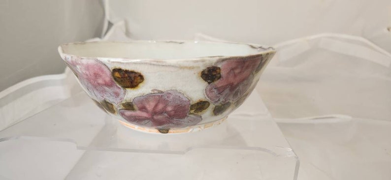 May include: A ceramic bowl with a white interior and exterior, decorated with pink and brown floral patterns. The bowl has a slightly uneven rim, giving it a rustic, handmade look. It is likely used for serving food.