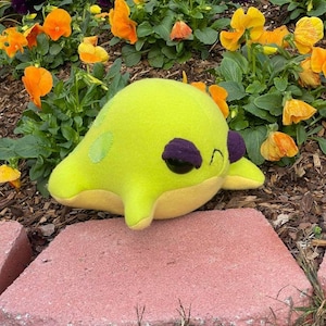 May include: A bright green plush toy shaped like a sea creature with large, black eyes and a purple stripe. The toy is sitting on a pink brick.