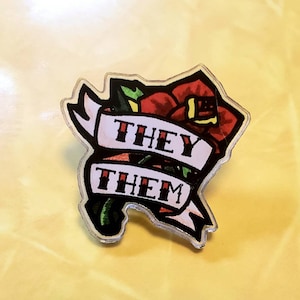 May include: A red rose with green leaves and a white banner that says "They Them" in black lettering. The rose and banner are on a clear acrylic background.