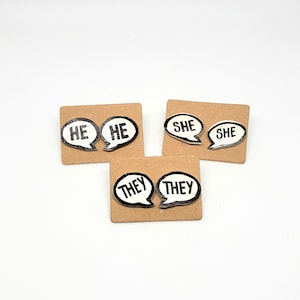 May include: Three pairs of earrings with speech bubble designs. The earrings say "HE", "SHE", and "THEY" in black text on a white background.