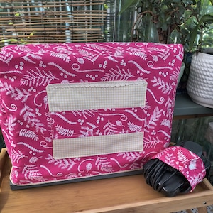 May include: A rectangular sewing machine cover in bright pink fabric with a white fern and floral pattern. It features a beige and white gingham pocket with lace trim. A matching fabric cord wrap with a white snap button is positioned nearby.