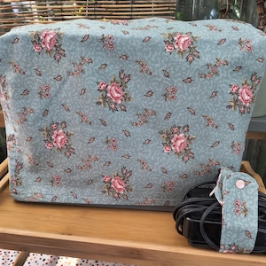Può includere: Una copertura in tessuto rettangolare con sfondo azzurro e un motivo ripetuto di rose rosa e piccole foglie verdi. Accanto alla copertura si trova un cavo di alimentazione nero avvolto da una fascetta in tessuto coordinata con bottone.