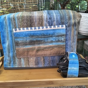 May include: A fabric sewing machine cover featuring a blue, brown, and grey marbled pattern with a rectangular front pocket. The pocket has a blue and white striped trim with lace detailing. A matching fabric cord wrap with a blue snap is included.