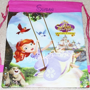 May include: Pink drawstring bag with a Sofia the First design. The bag features a picture of Princess Sofia on a swing with a castle in the background. The text "Susan" is printed on the top of the bag.