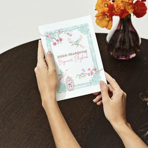 May include: A white Mahjong Beginner Playbook for 2026 is held by a person. The cover features a teal border with floral and bird illustrations. The book is on a dark brown table, with a vase of orange flowers in the background.