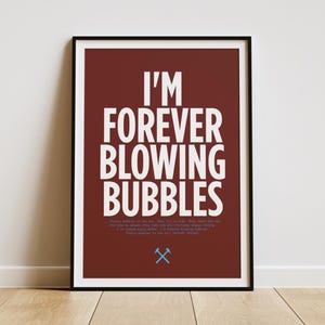 Op de afbeelding: Een ingelijste kunstdruk met de tekst "I'M FOREVER BLOWING BUBBLES" in wit op een bordeauxrode achtergrond. De print bevat een kleine afbeelding en extra tekst onderaan. De lijst is zwart en het kunstwerk wordt tegen een witte muur getoond.