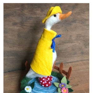 May include: A white felt goose wearing a yellow raincoat and hat, standing in a felt pond with red and white polka dot boots. The goose is surrounded by green felt grass and flowers.