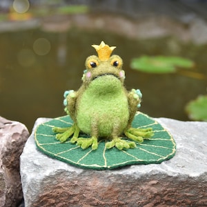 Könnte beinhalten: Ein grüner Filz-Frosch mit einer goldenen Krone sitzt auf einem grünen Filz-Seerose. Der Frosch sitzt auf einem Stein in der Nähe eines Teichs.