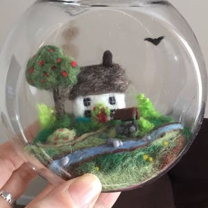 May include: A miniature felt scene of a white cottage with a brown roof, a green tree with red apples, a small stream, and a black bat flying overhead, all enclosed in a clear glass jar with a black lid.