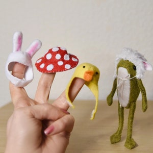 Peut inclure: Trois marionnettes à doigts, un lapin blanc, un champignon rouge et blanc et un canard jaune, sont portées sur les doigts d'une personne. Une marionnette de grenouille verte se tient sur une surface en bois.