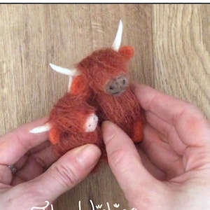 May include: Two small, hand-felted Highland Cow figurines. The cows are brown and have white horns. The smaller cow is nestled against the larger cow. The text "The Wishing Shed" is written in white on a wooden background.