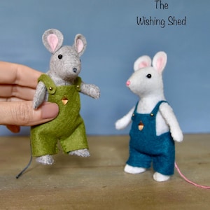Könnte beinhalten: Zwei Filzmäuse mit Latzhosen. Eine Maus ist grau und trägt eine grüne Latzhose, die von einer Hand gehalten wird. Die andere Maus ist weiß mit blauer Latzhose. Beide haben rosa Ohren und Nasen. Der Text "The Wishing Shed" steht über den Mäusen.