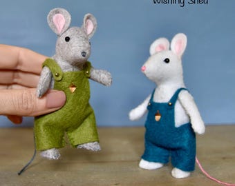 May include: Two felt mice wearing overalls. One mouse is gray and wearing green overalls, held by a hand. The other mouse is white with blue overalls. Both have pink ears and noses. The text "The Wishing Shed" is above the mice.