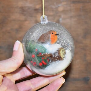 May include: A clear glass Christmas ornament with a felt robin perched on a branch with red berries and snow inside.