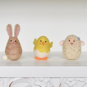 May include: Three felt animal figurines: a brown bunny, a yellow chick, and a white sheep. The bunny has long ears and a white tail. The chick has orange feet and a white egg shell. The sheep has a blue flower on its head.