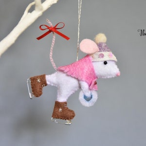 Puede incluir: Adorno de ratón de fieltro blanco con un gorro rosa y un abrigo rosa, patinando sobre hielo con un círculo de fieltro azul en la mano. El ratón tiene un lazo rojo en la cola. El adorno está colgado de una cuerda blanca.