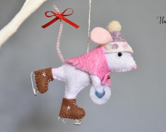 Op de afbeelding: Witte vilt muis ornament met een roze hoed en een roze jas, schaatsen met een blauwe vilt cirkel in zijn hand. De muis heeft een rode strik op zijn staart. Het ornament hangt aan een wit touwtje.