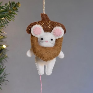 Può includere: Un ornamento in feltro a forma di topo bianco con orecchie e naso rosa, che indossa un cappello a forma di ghianda marrone e tiene in mano un guscio di ghianda in feltro marrone. Ha occhi a perla neri, un filo argentato e un filo rosa.