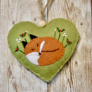 Könnte beinhalten: Herzförmiger grüner Filzschmuck mit einem schlafenden Fuchs-Design. Der Fuchs ist orange und weiß, umgeben von gestickten Blumen und Beeren. Eine Holzperle und eine Schnurschlaufe sind zum Aufhängen befestigt.