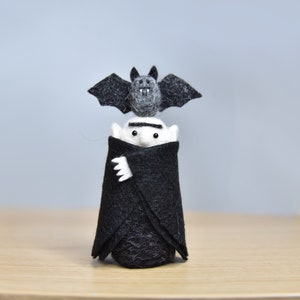 May include: A small felt figurine of a vampire wearing a black cape with a bat perched on its head.