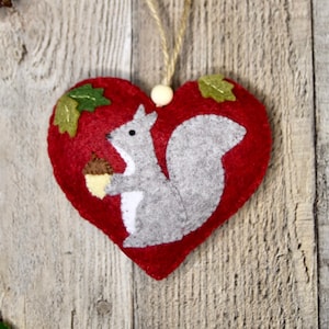 Puede incluir: Adorno en forma de corazón rojo de fieltro con una ardilla gris sosteniendo una bellota. Detalles de hojas verdes adornan la parte superior. Una cuerda beige está unida para colgar.