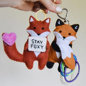 May include: Two felt fox keychains. The first is a rust-coloured fox with a white chest, black eyes, and a pink heart on its tail, holding a white sign that reads STAY FOXY. The second is a brown and white fox with black ears and paws.