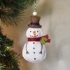 Pode incluir: Enfeite de Natal de boneco de neve feito à mão. O boneco de neve branco tem um nariz de cenoura, olhos e boca de pontos pretos e um cachecol vermelho. Ele usa uma cartola marrom com uma faixa azul e um trevo verde. O enfeite é pendurado por uma corda verde e branca.