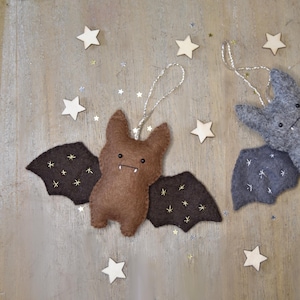 May include: Two felt bat ornaments, one brown and one gray, with embroidered wings and small white fangs. Each bat has a string for hanging. Wooden star decorations and glitter are scattered around the bats.
