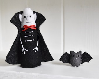 May include: A felt vampire doll with a white face, black cape, and red ribbon bow tie, accompanied by a small gray felt bat. The vampire features embroidered white details on the black coat and simple facial features with two small fangs.