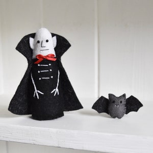 Puede incluir: Una muñeca de vampiro de fieltro con una capa negra y una pajarita roja se encuentra junto a una muñeca de murciélago de fieltro. Ambas muñecas tienen rasgos faciales bordados.