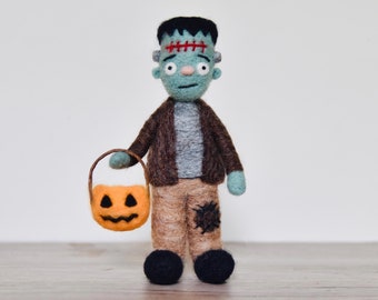 May include: A green felt Frankenstein monster doll holding a small orange pumpkin bucket. The doll is wearing a brown jacket and tan pants.