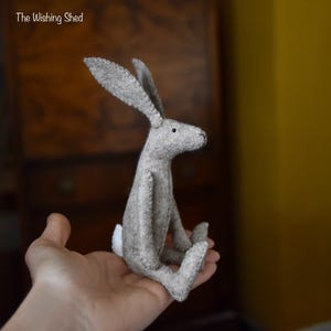 Puede incluir: Un conejo de fieltro gris con orejas largas y una pequeña cola blanca. El conejito hecho a mano está sentado erguido, con las piernas extendidas. El texto "The Wishing Shed" está en la esquina superior izquierda.