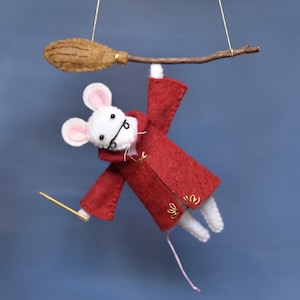 May include: A charming felt mouse in a red robe, clutching a wand, hanging from a miniature broom. The mouse has white fur, pink ears, and tiny spectacles. The broom is brown, set against a plain blue backdrop.