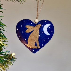 Peut inclure: Ornement de Noël en forme de cœur, avec un fond bleu foncé. Il présente une silhouette de lièvre beige, des étoiles blanches et un croissant de lune. L'ornement est suspendu par une ficelle et une perle en bois.