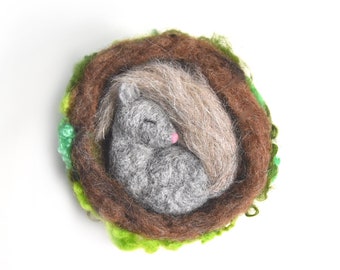 May include: A needle-felted brooch featuring a small gray squirrel curled up inside a brown nest. The squirrel has a pink nose and a long, textured gray tail wrapped around its body. The nest is surrounded by bright green felted accents.
