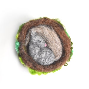May include: A needle felted squirrel sleeping in a brown and green nest. The squirrel is grey with a pink nose and a brown tail.