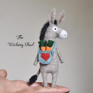 May include: A gray felt donkey with black mane and tail, holding a bunch of orange carrots in a blue felt pocket with a red heart. The donkey is standing on a hand.