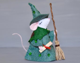 May include: A felt mouse dressed as a witch with a green hat, green robe, and a brown broom. The mouse is holding a scroll in its hand.